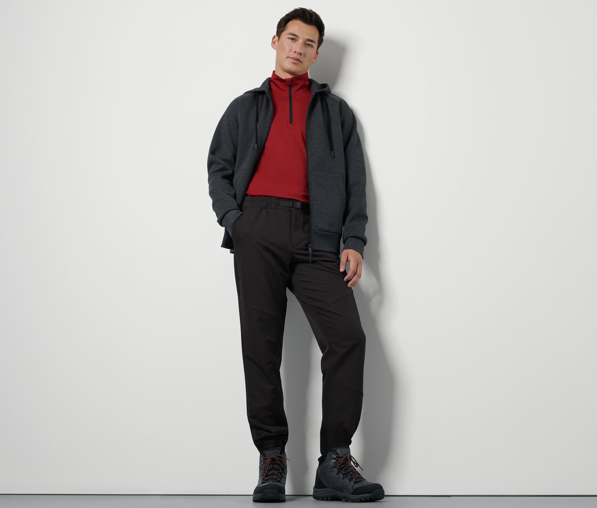 Muž stojí a má na sobě termokalhoty s páskem, mikinu na zip a turistické boty.