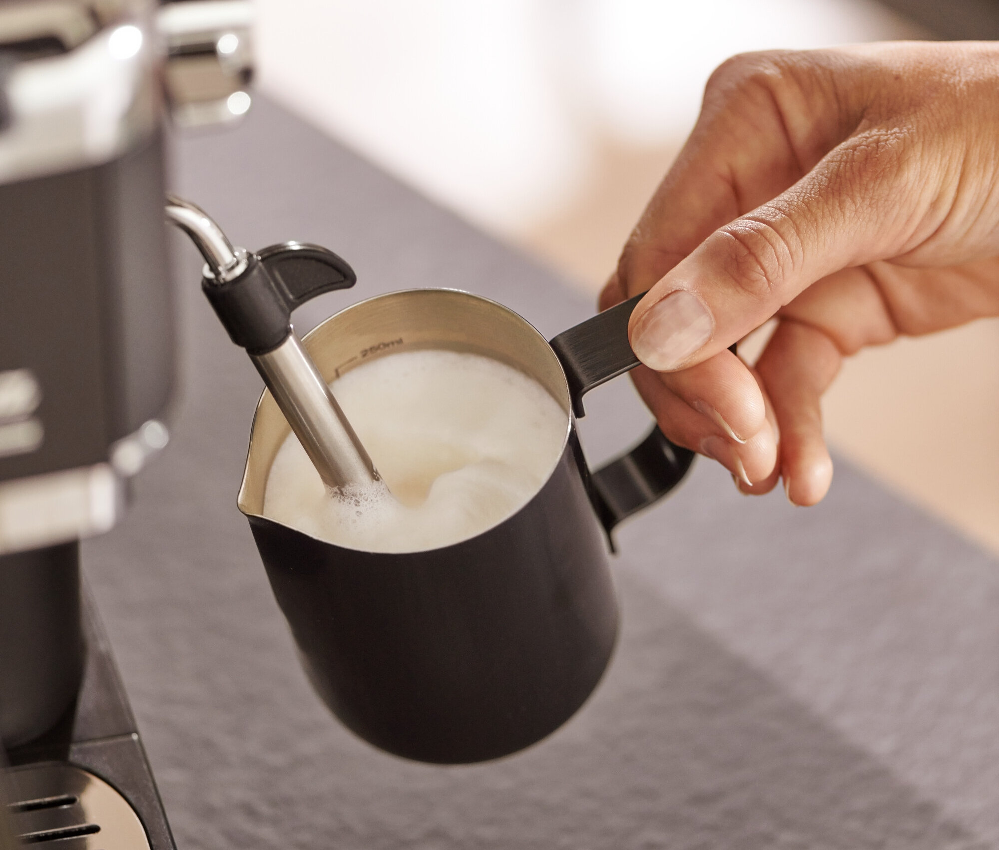 Osoba napěňuje mléko v černé mlékovce pomocí kávovaru.