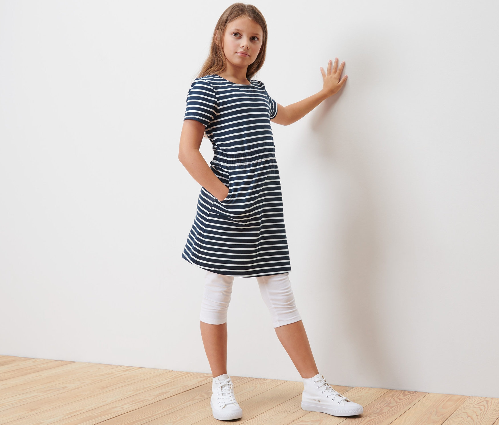 Dívka opřená o stěnu má na sobě dívčí žerzejové šaty s modrými a bílými pruhy.