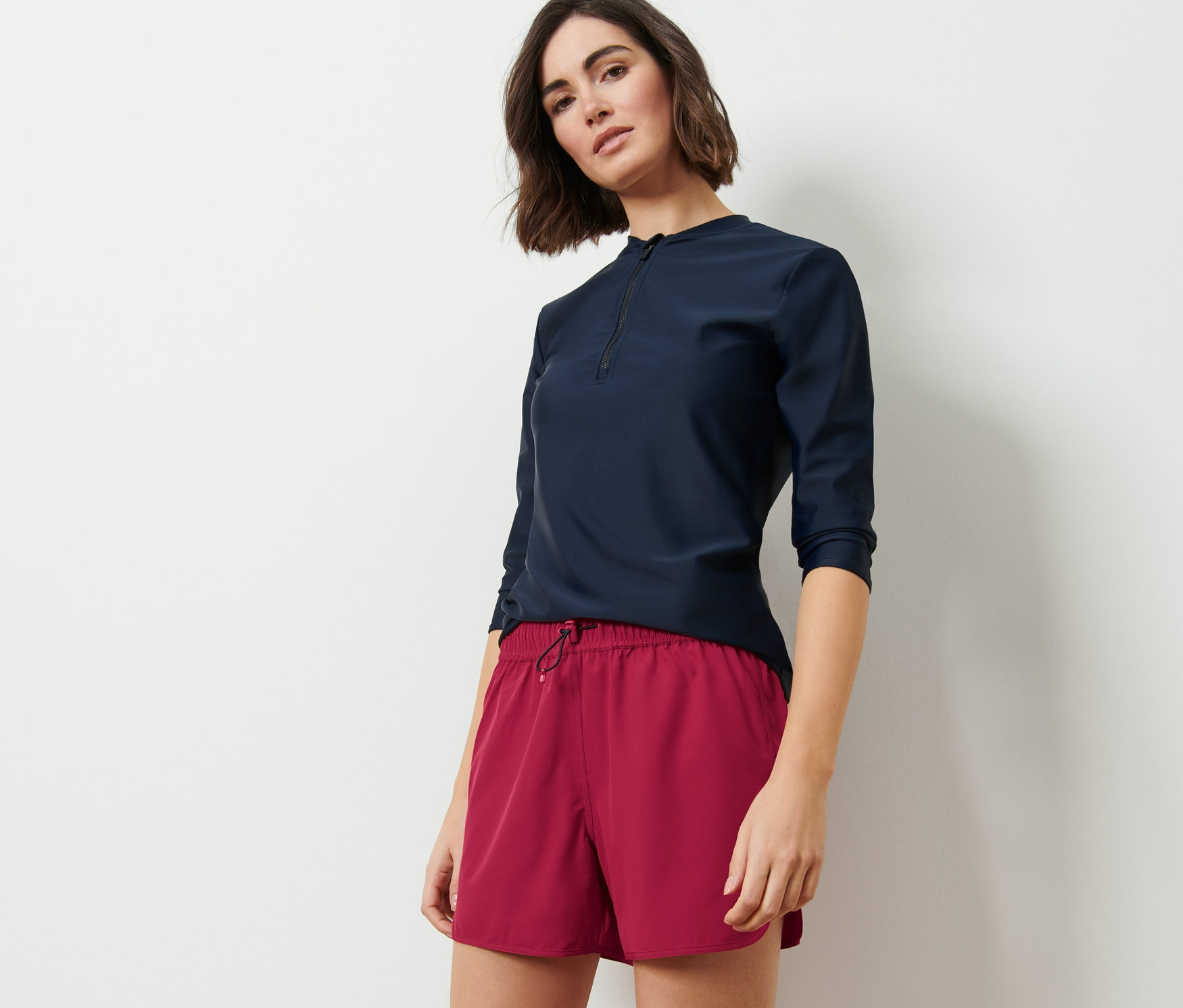 Žena pózuje v tmavě modrém topu s tříčtvrtečním rukávem a bobulových koupacích šortkách.