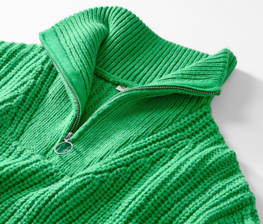 Detail zeleného svetru troyer z hrubého úpletu s částečně rozepnutým zipem.