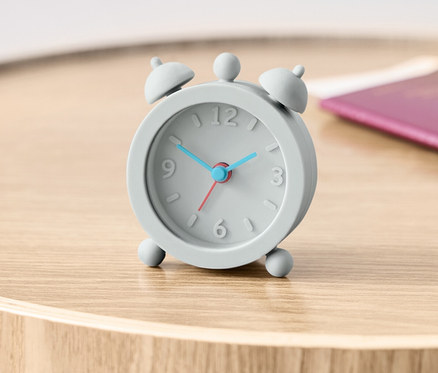 Šedý cestovní budík na dřevěném stolku vedle pasu.