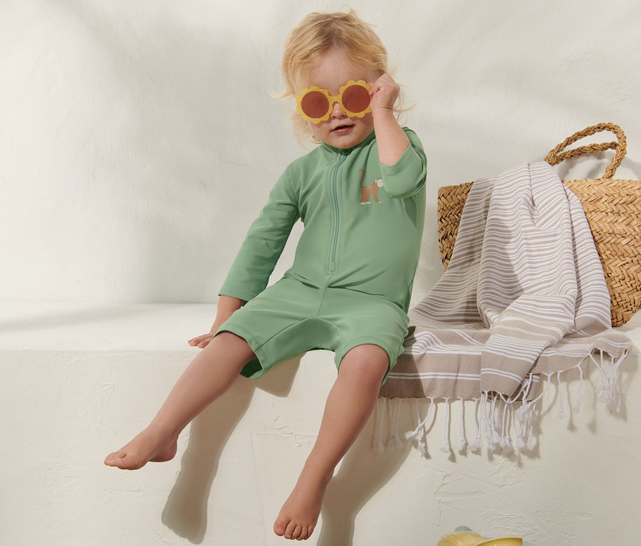 Dítě sedí a má na sobě zelený overal s UV ochranou 80 a sluneční brýle.