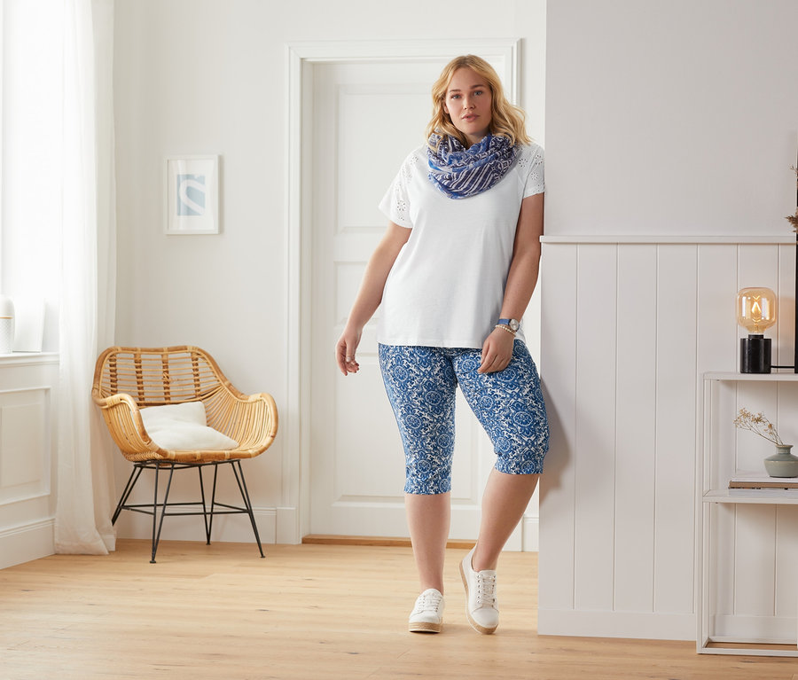 Žena opřená o stěnu má na sobě bílé tričko s výšivkou a modré 3/4 strečové kalhoty s potiskem.