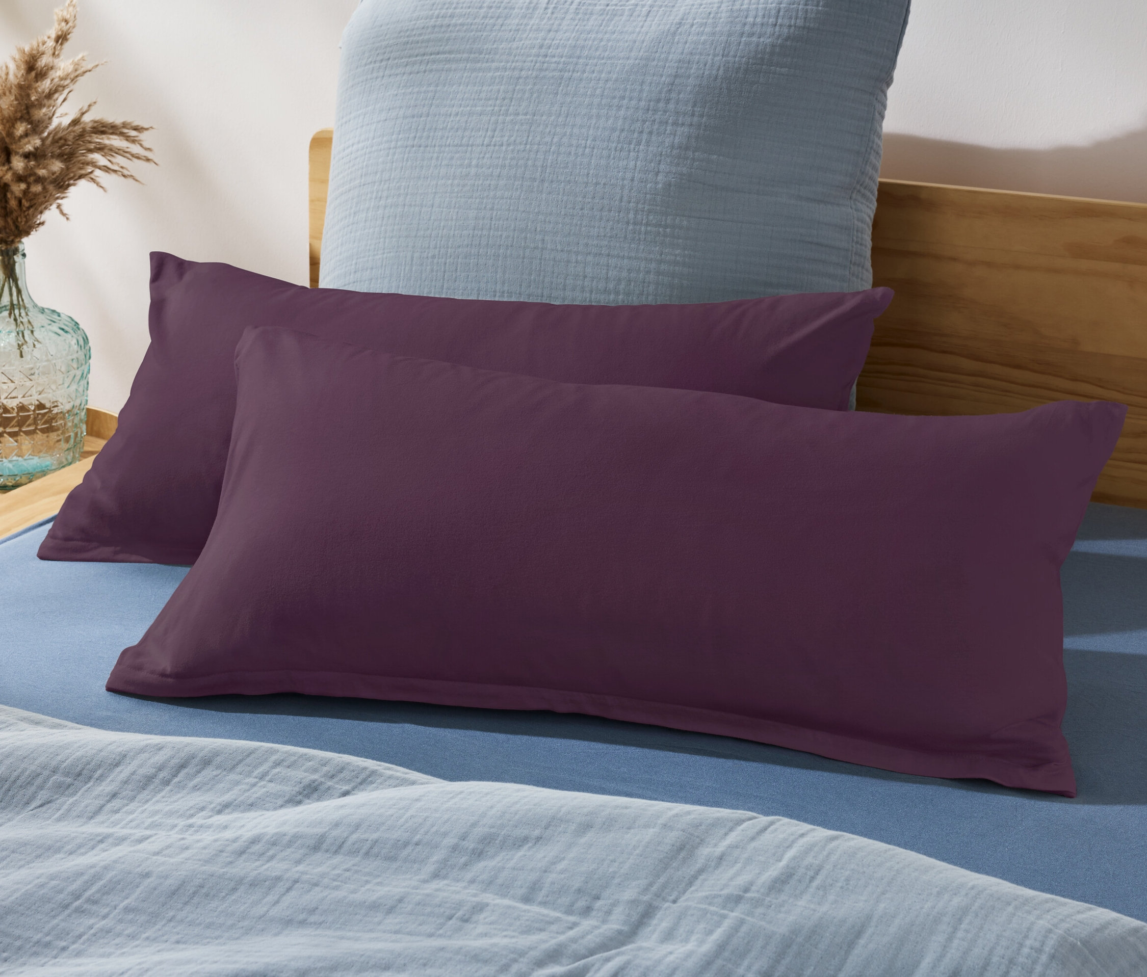 Dva fialové polštáře na posteli s modrým prostěradlem a modrým polštářem v pozadí.