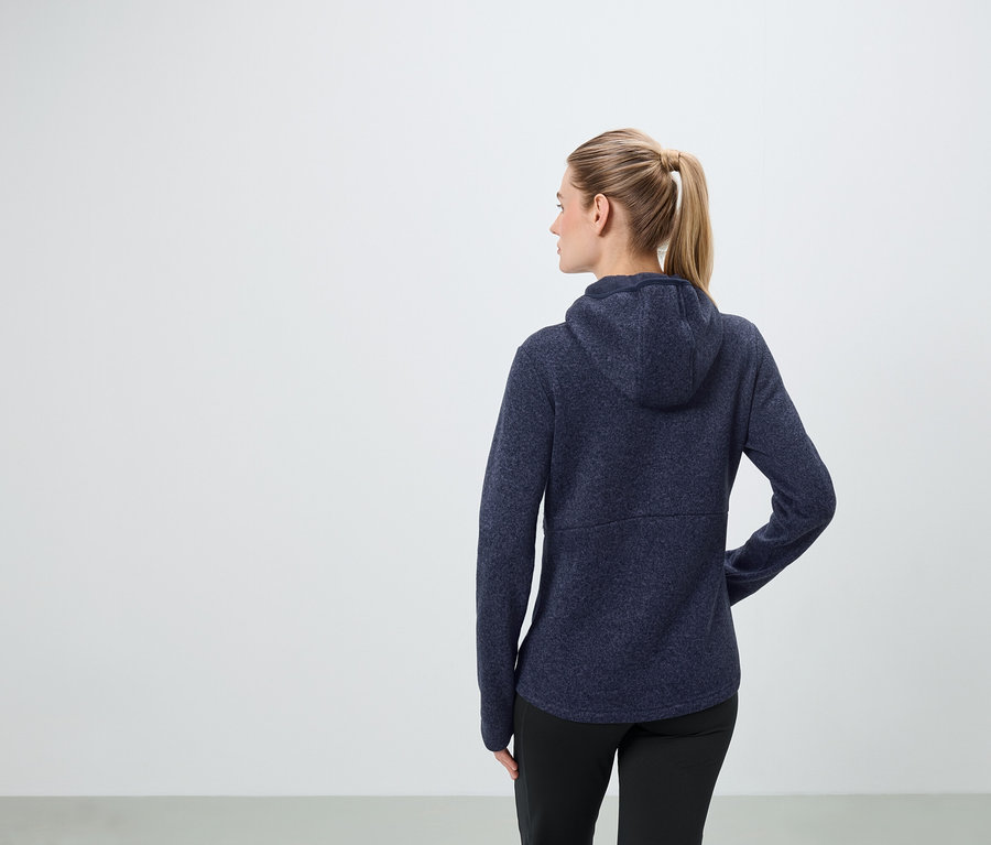 Žena s blond vlasy staženými do culíku má na sobě tmavě modrou mikinu s kapucí a černé kalhoty.