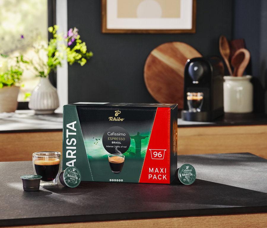 Na stole stojí balení kapslí Cafissimo BARISTA Espresso Brasil, sklenice s espressem a kávovar.