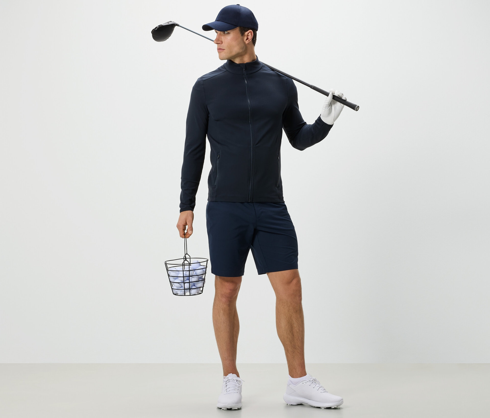 Muž v tmavě modré funkční bundě drží koš s golfovými míčky a golfovou hůl.