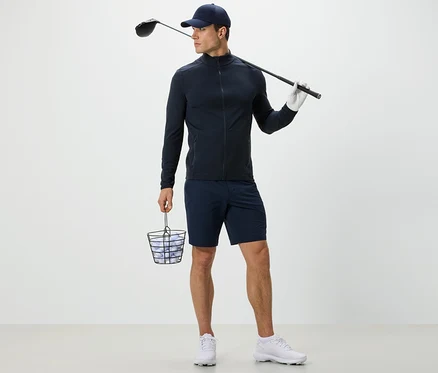 Muž v tmavě modré funkční bundě drží koš s golfovými míčky a golfovou hůl.