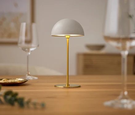 Na dřevěném stole stojí bílá lampa, sklenice a talíř s jídlem.