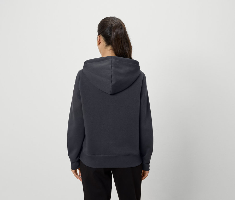 Zezadu stojící žena má na sobě tmavě šedou mikinu s kapucí a černé kalhoty.