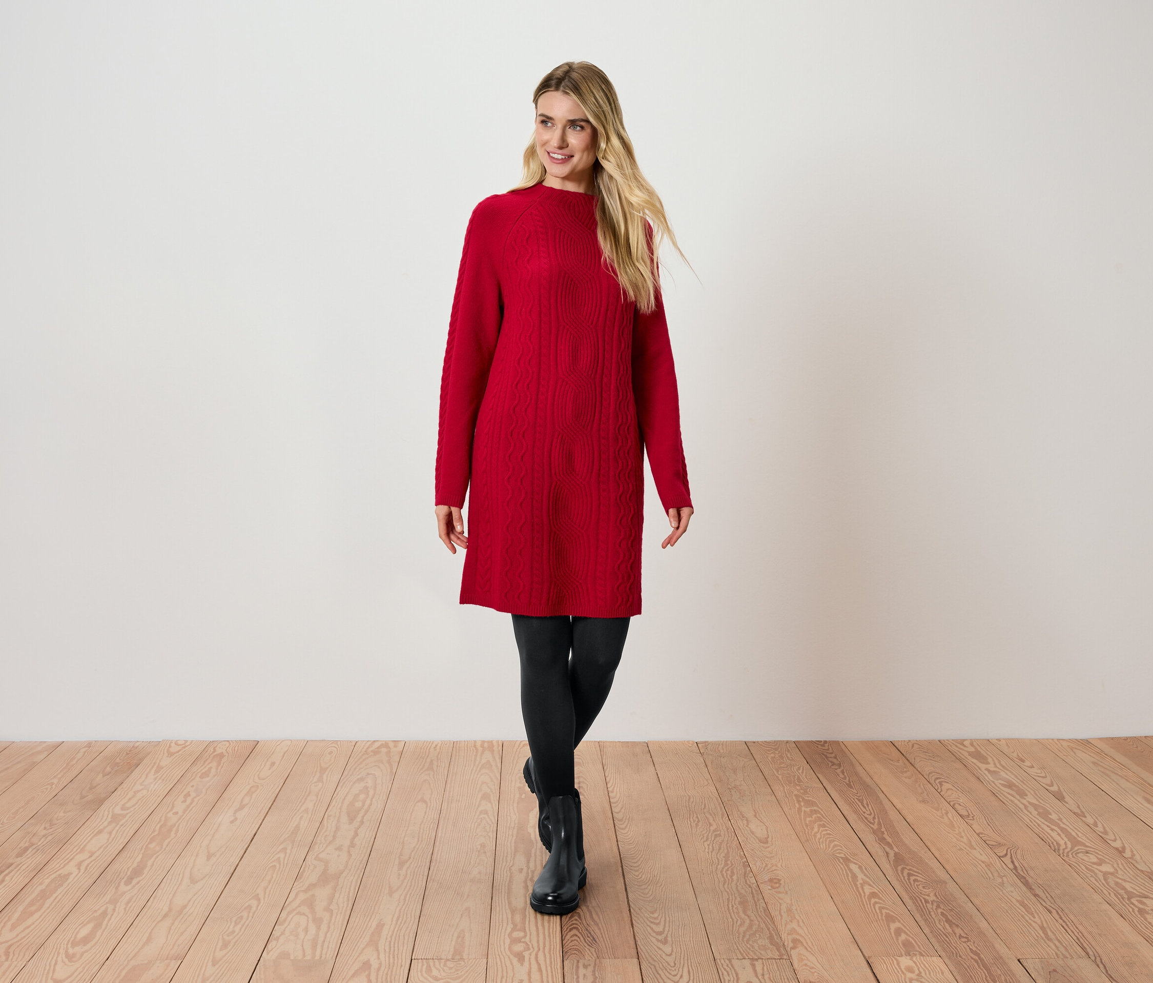 Blondýna v červených pletených šatech, černých legínách a černých botách stojí na dřevěné podlaze.