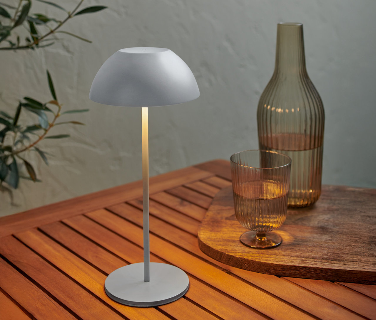 Šedá lampa stojí na dřevěném stole vedle láhve a sklenice vody.