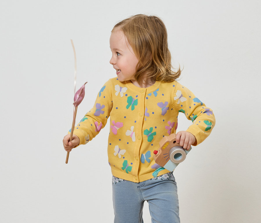 Holčička ve žlutém svetru s motýly drží hračku fotoaparátu a květinu.