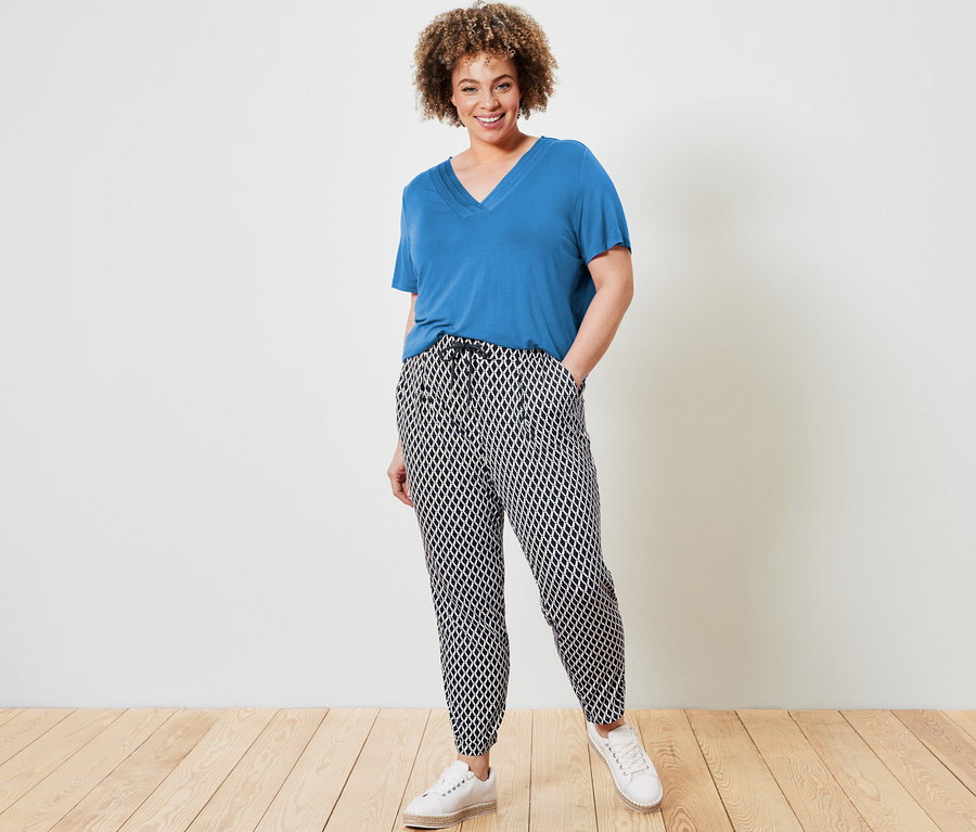 Žena pózuje v kobaltově modrém tričku s detailem skladů a vzorovaných žerzejových kalhotách.