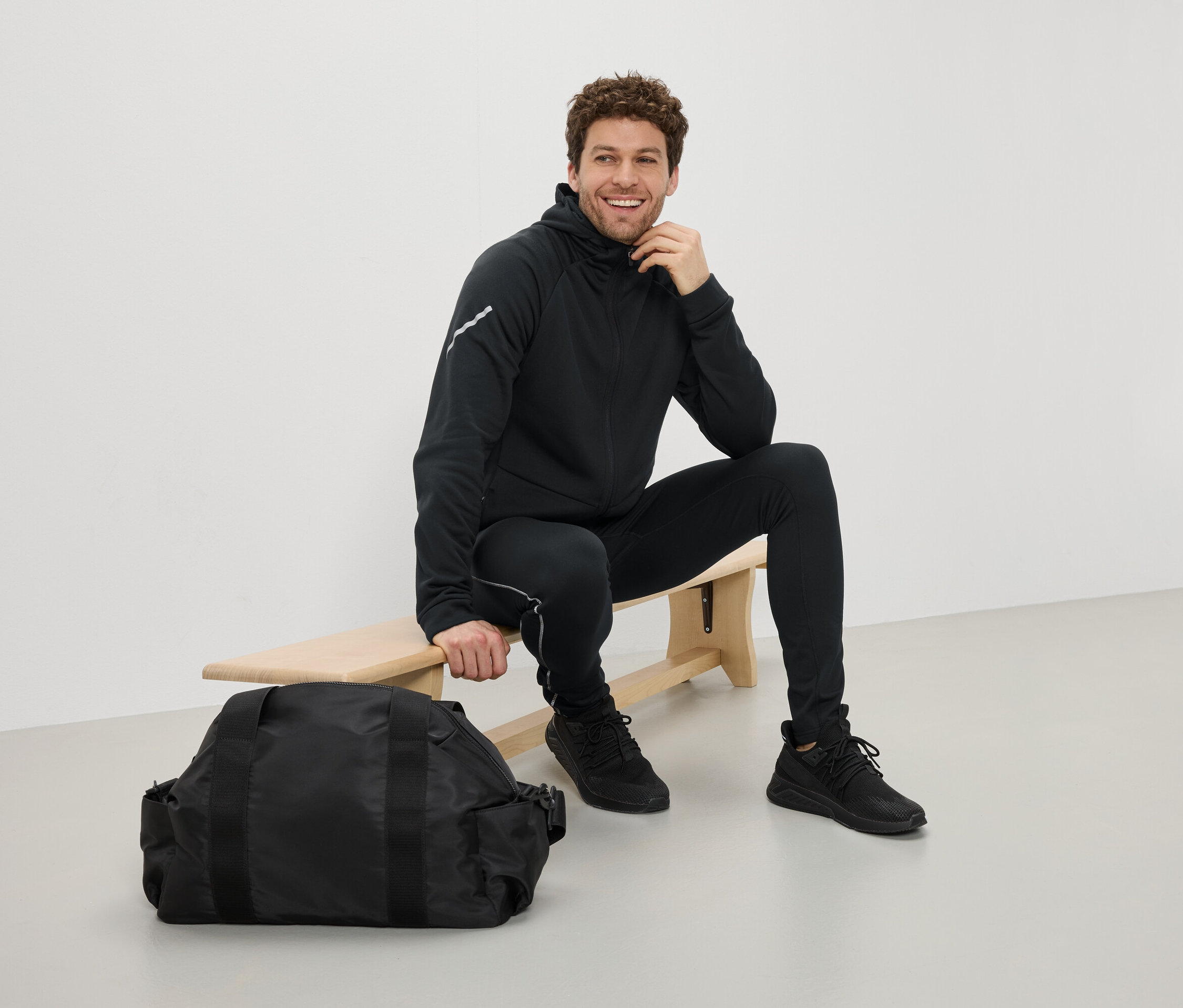 Muž sedí na lavici v černém sportovním oblečení vedle černé sportovní tašky.