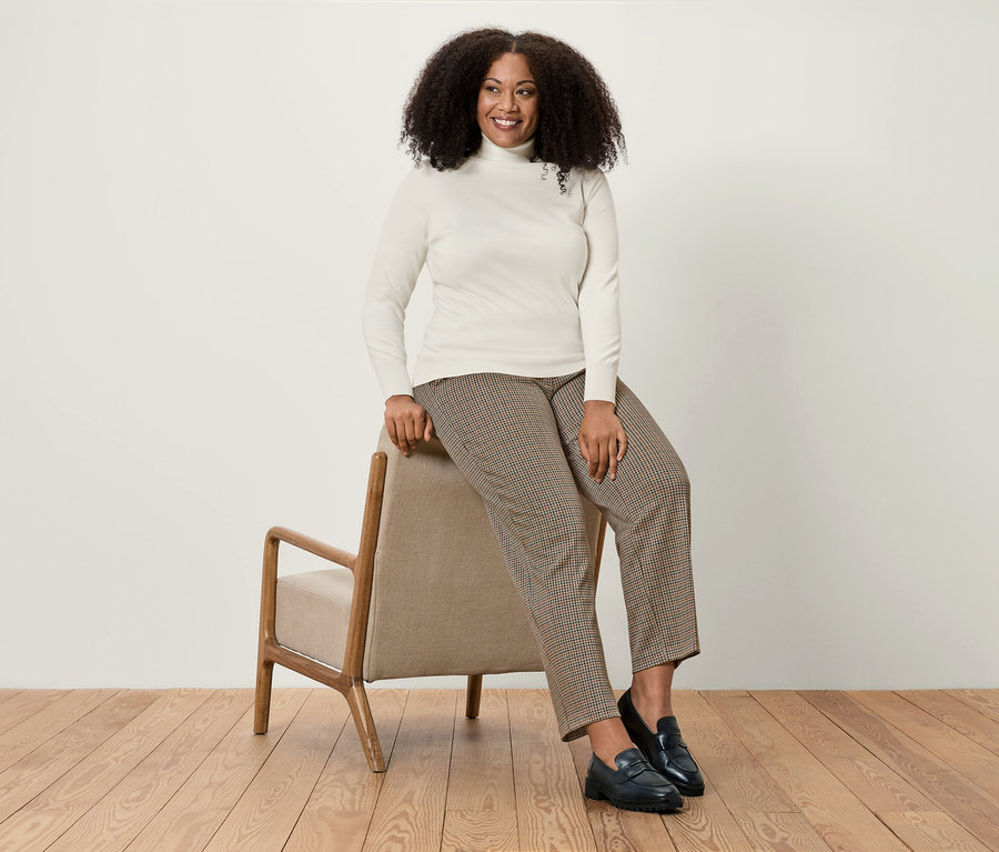 Žena sedí na béžovém křesle, má na sobě bílý rolák, hnědé kostkované kalhoty a černé mokasíny.