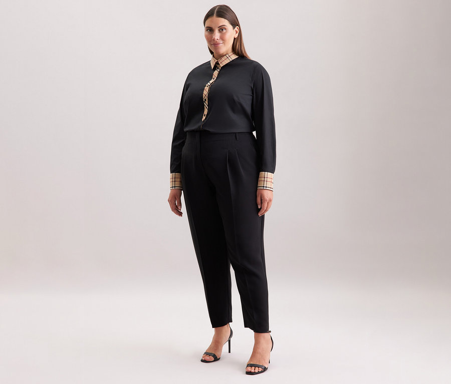 Žena má na sobě černé kalhoty a popelínovou halenku Seidensticker s kostkovaným detailem a černé boty na vysokém podpatku.