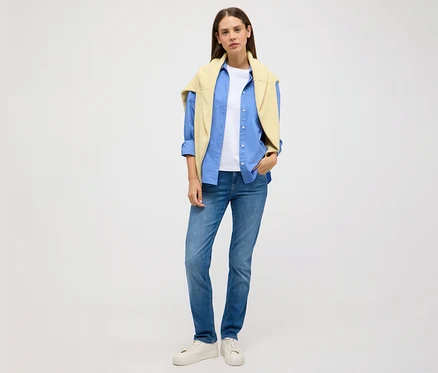 Žena stojící na světlém pozadí má na sobě džíny Angels »Cici Push-Up« slim fit, modrou košili, bílé tričko a žlutý svetr.
