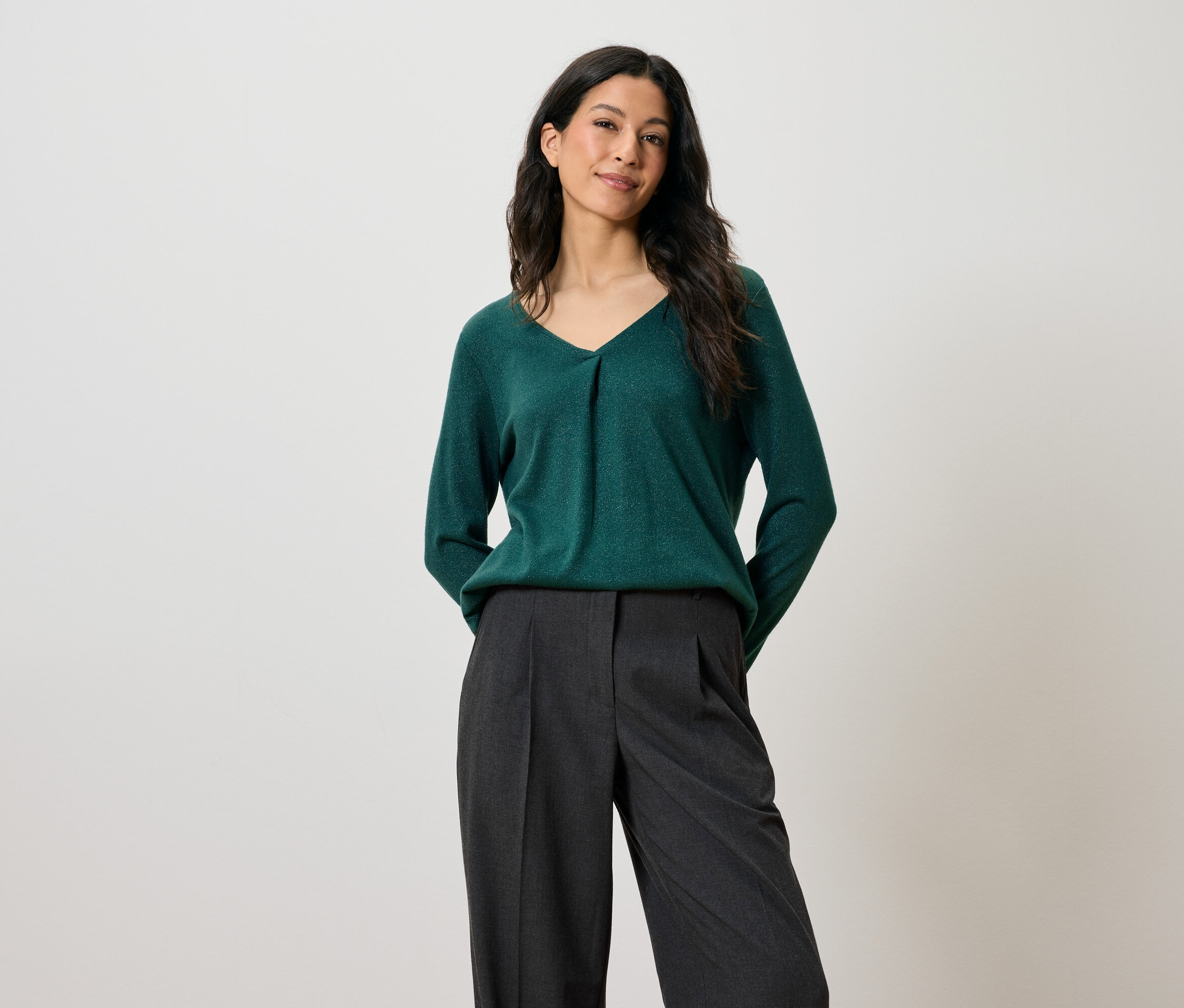 Žena s tmavými vlasy pózuje v zeleném svetru a šedých kalhotách.
