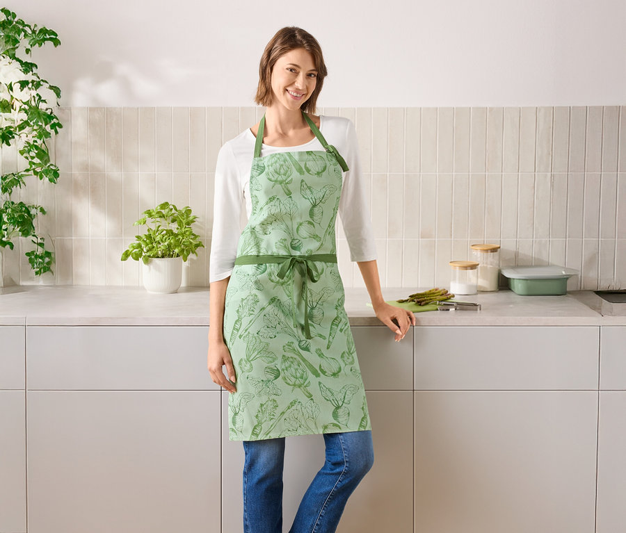 Žena stojí v kuchyni a má na sobě zelenou zástěru se zeleninou.