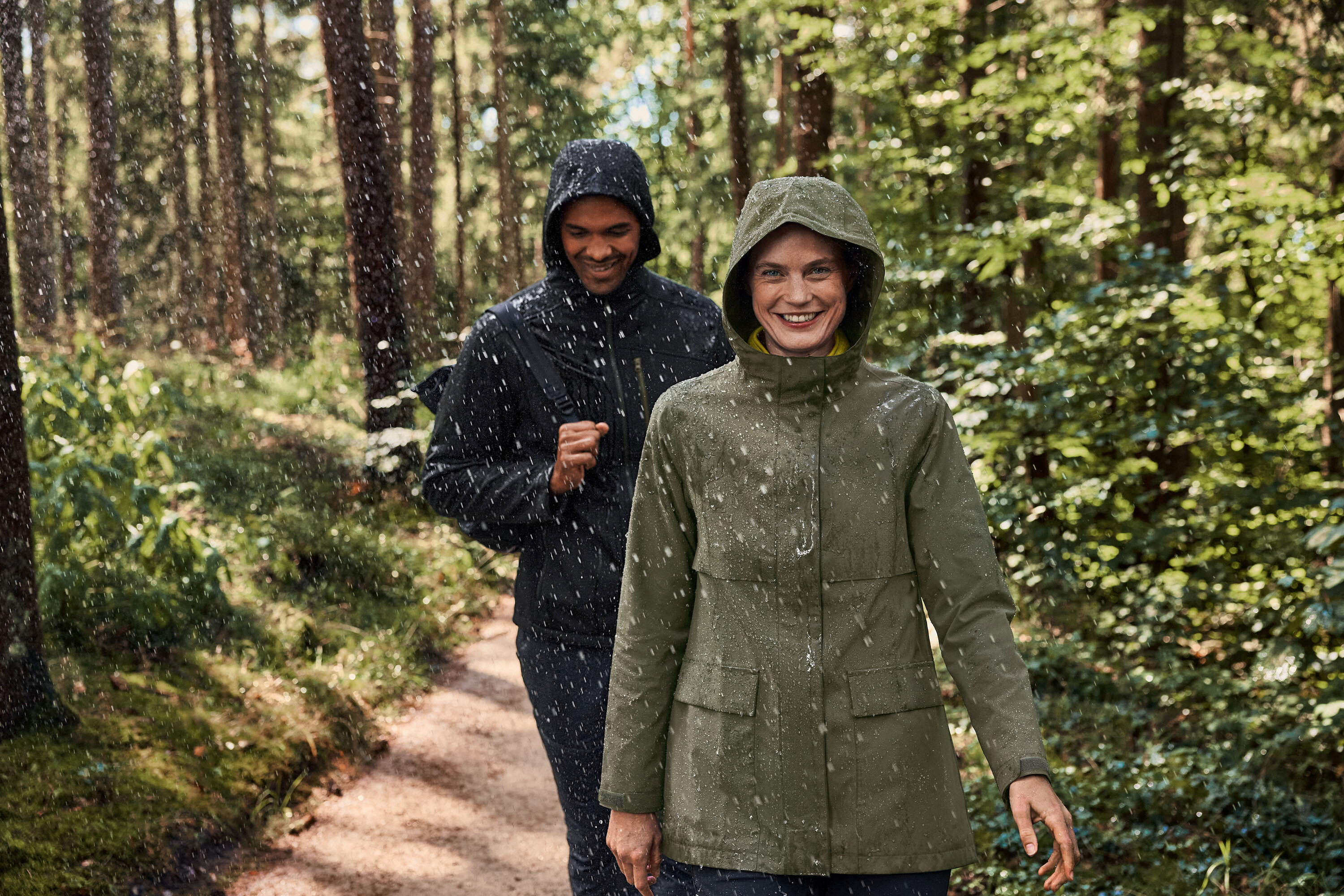 Dva lidé kráčejí v dešti lesem. Žena má na sobě fleecový svetr troyer, khaki/antracitová.