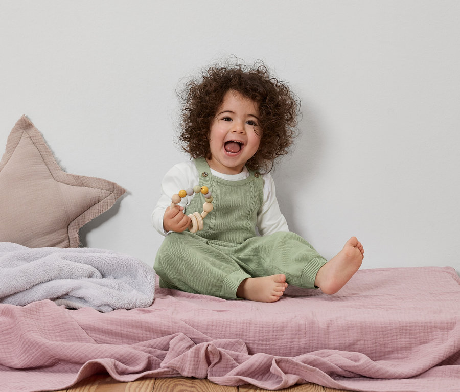 Usmívající se batole s kudrnatými vlasy sedí na růžové hrací dece a drží dřevěnou hračku.