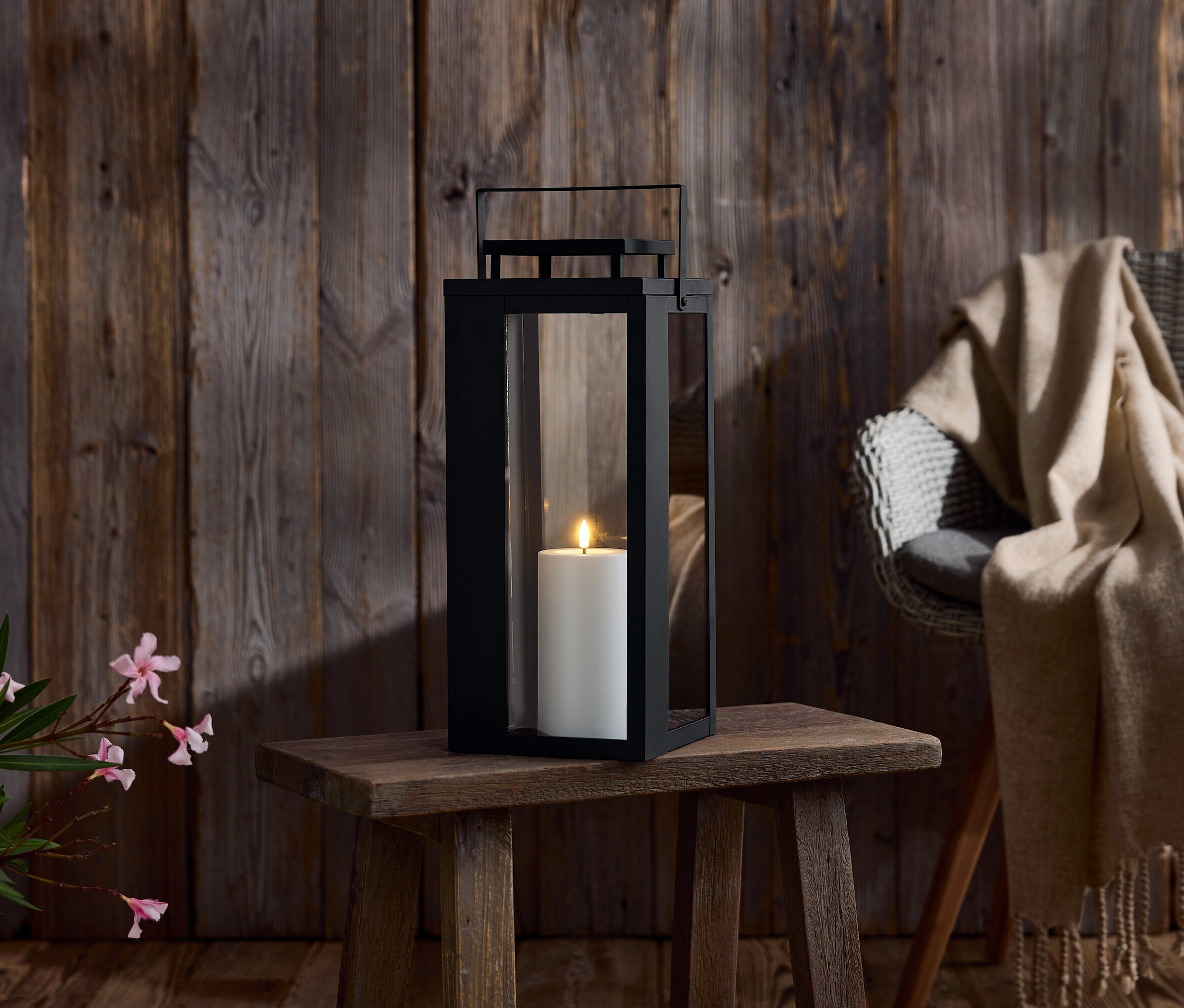 Černá lucerna se svíčkou na dřevěném stole. V pozadí dřevěná stěna a židle s dekou.