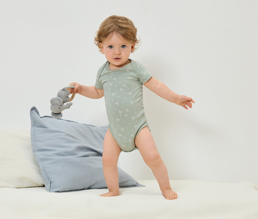 Batole v zeleném body s potiskem srdíček stojí na posteli a drží šedou hračku slona.