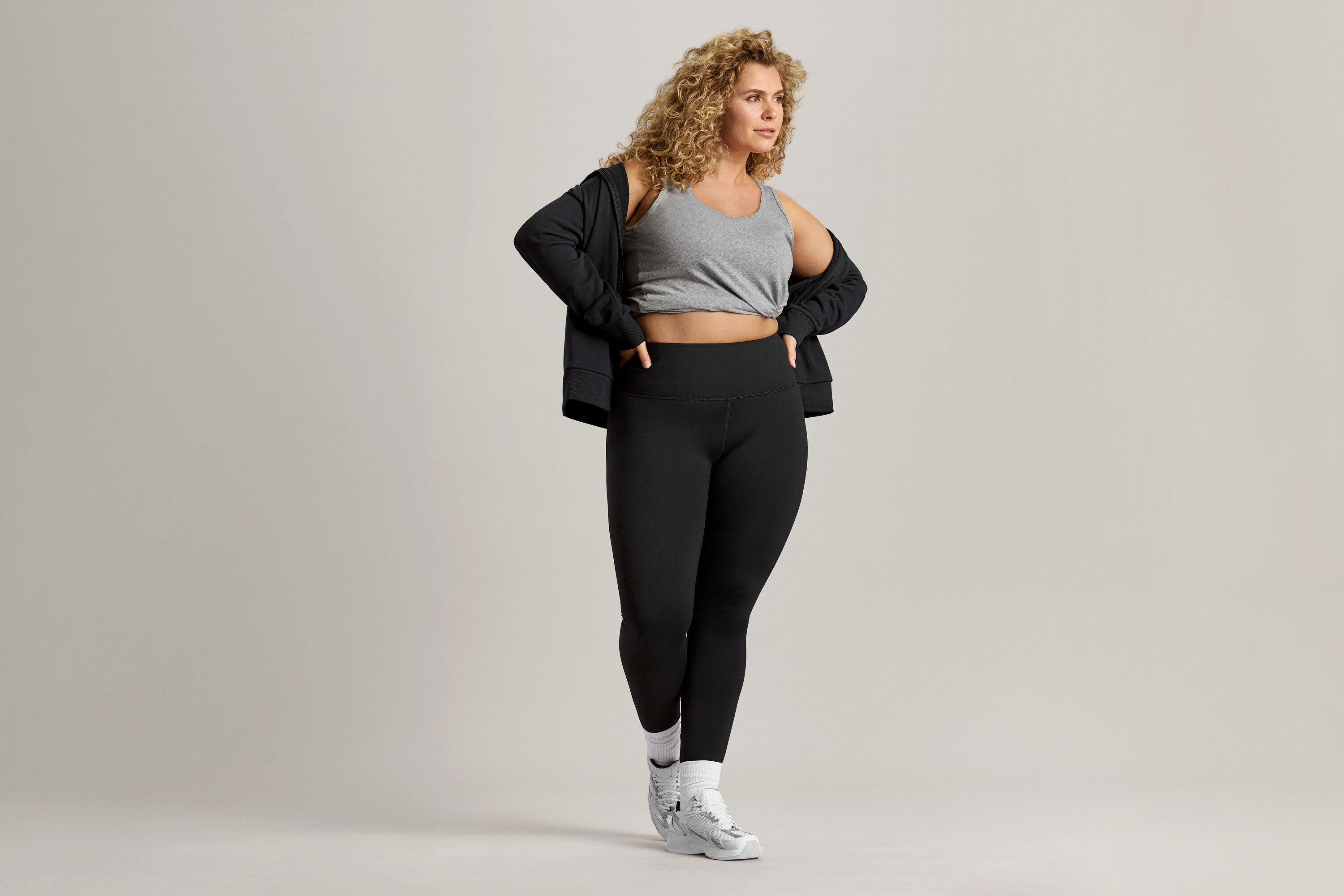 Žena s kudrnatými vlasy má na sobě šedé tílko, černou bundu a černé legíny.