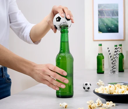 Osoba otevírá zelenou láhev piva otvírákem na láhve ve tvaru fotbalového míče. Na stole jsou popcorn a další láhve.