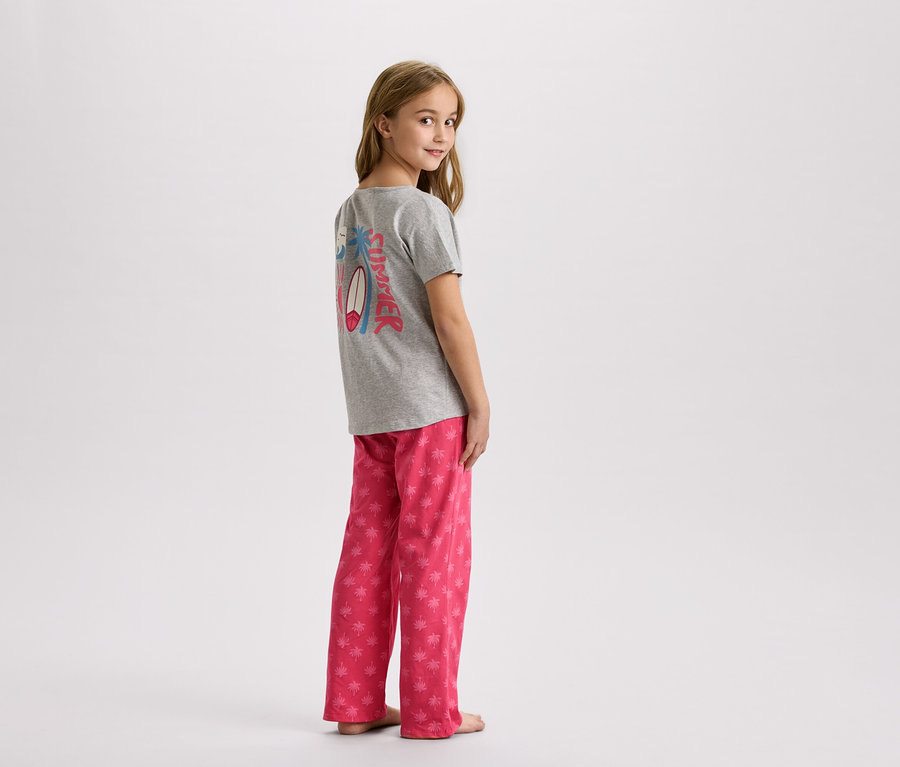 Dívka stojí v šedém tričku s letním designem a růžových kalhotách s palmami.