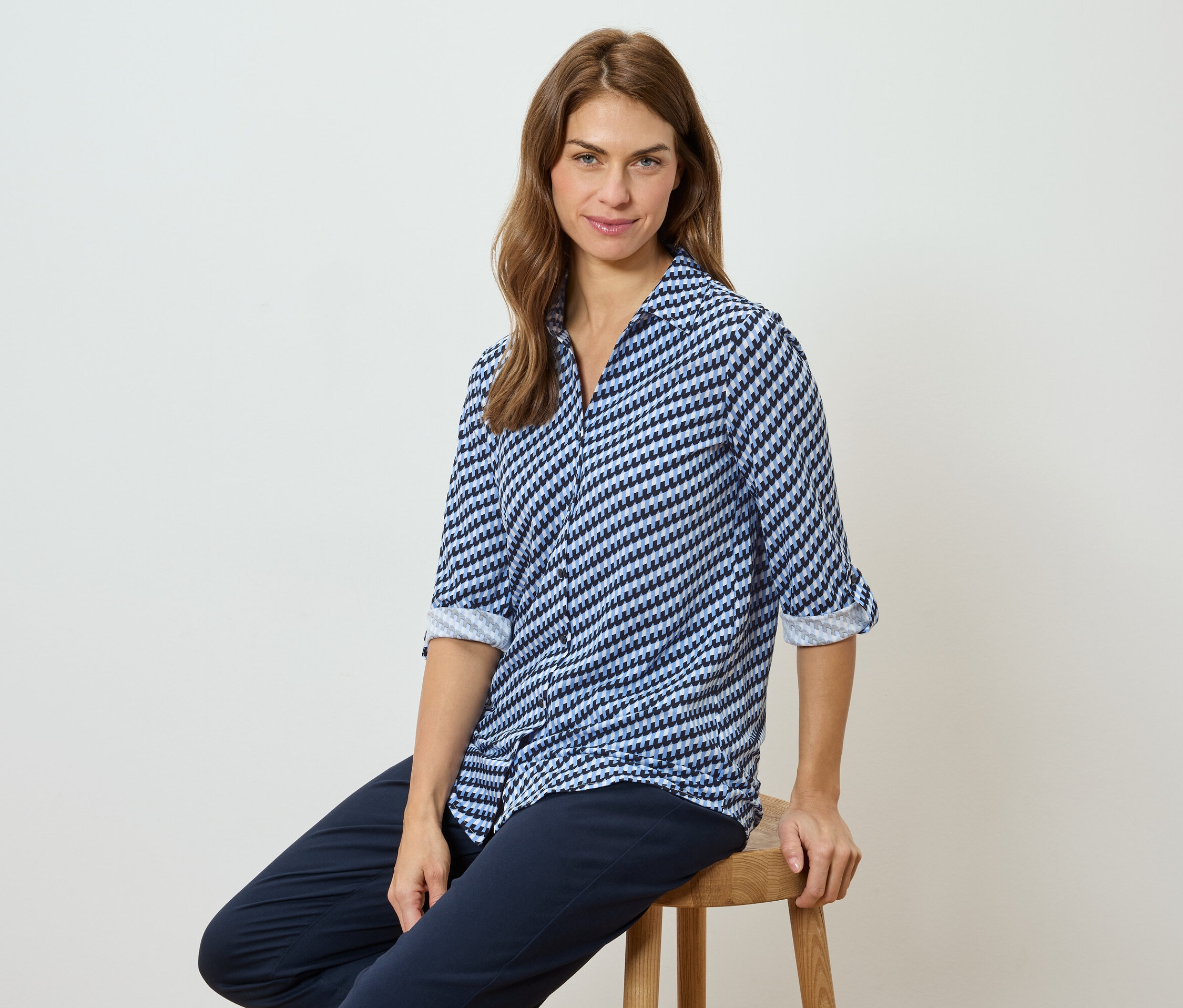 Žena sedí na stoličce a má na sobě modro-bílou vzorovanou halenku a tmavé kalhoty.