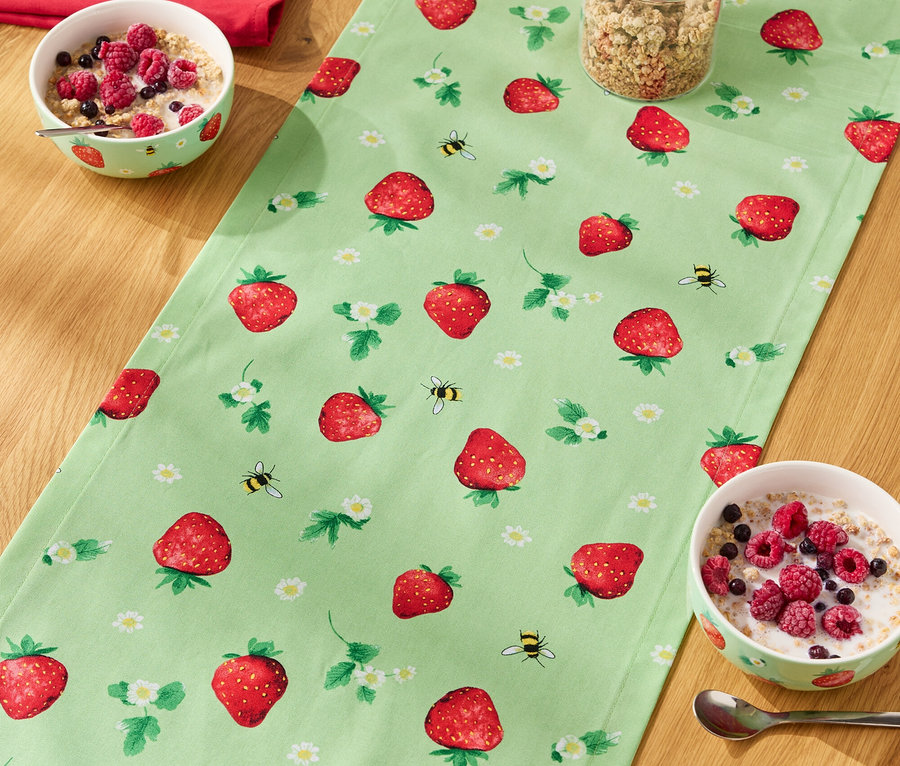 Na dřevěném stole leží běhoun se vzorem jahod, na něm misky s cereáliemi, sklenice müsli a láhev mléka.