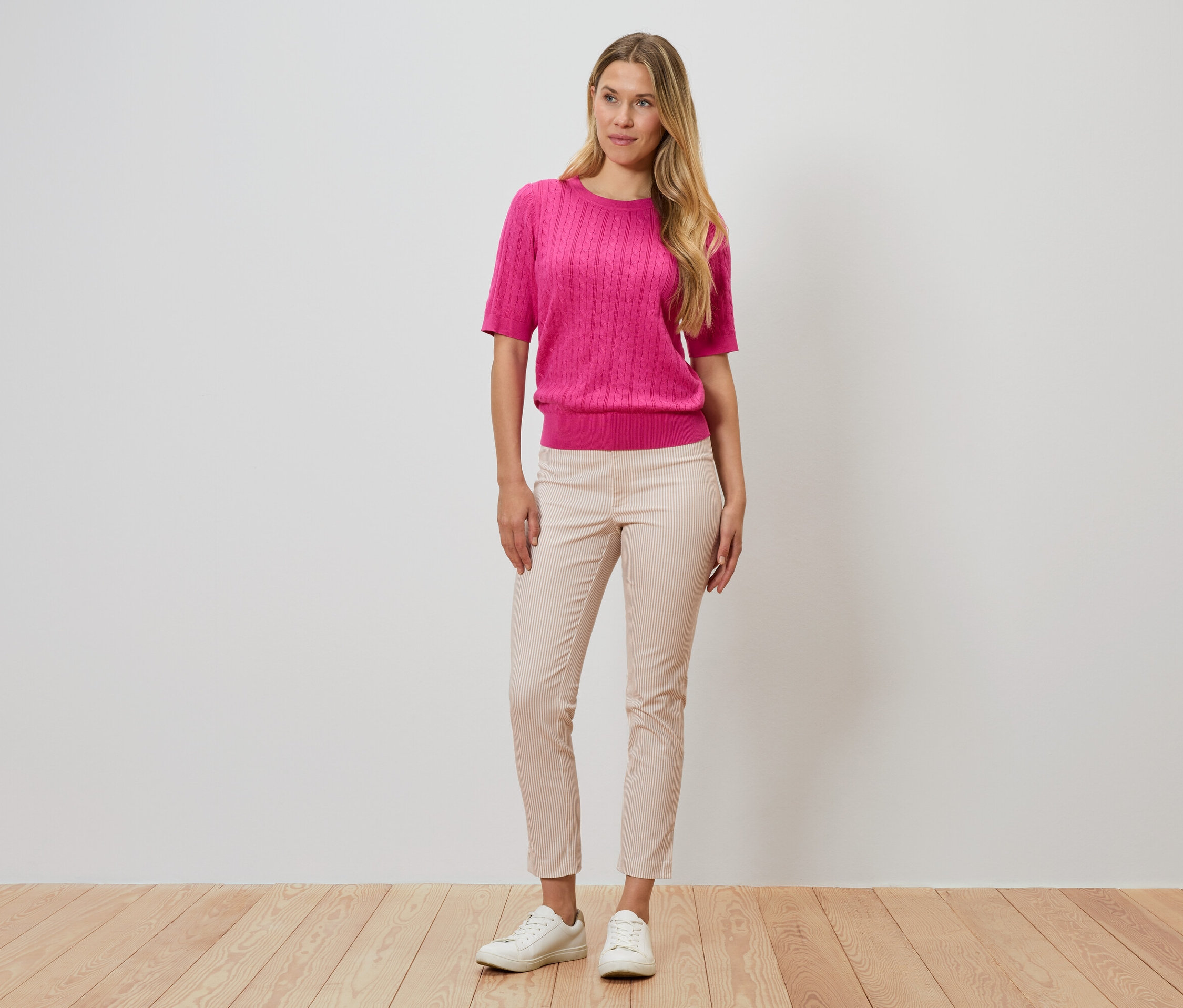 Žena stojí před zdí, má na sobě růžový svetr, pruhované kalhoty a bílé boty.