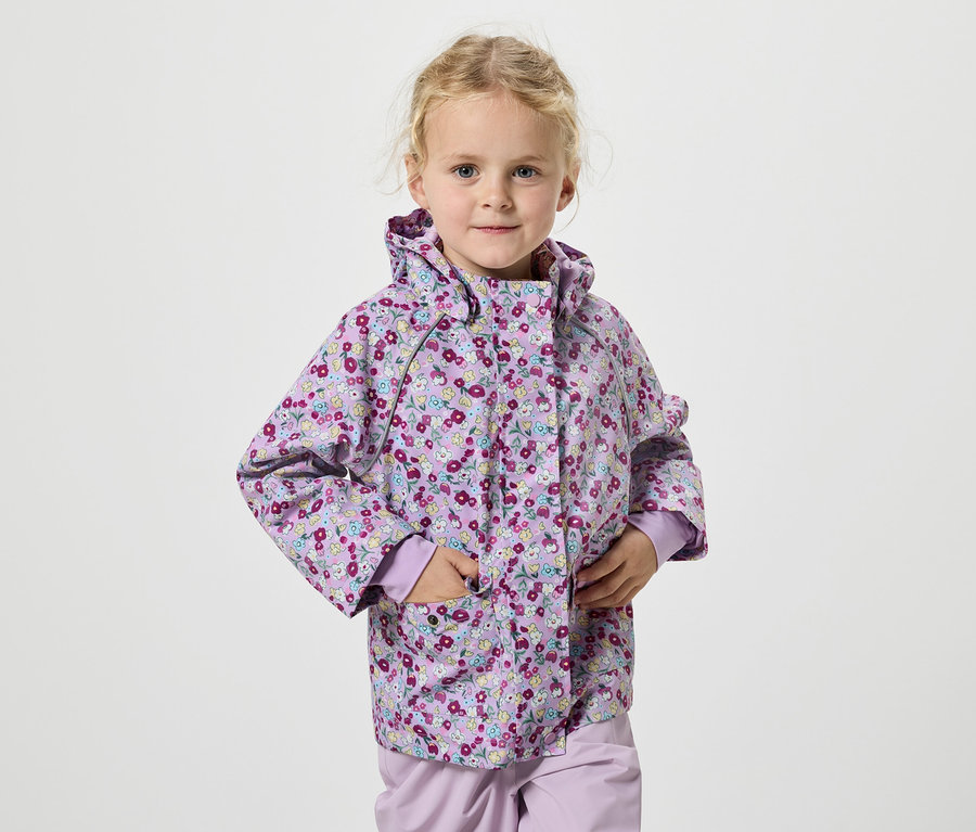 Dívka s blond vlasy v lila květinové bundě stojí na bílém pozadí.