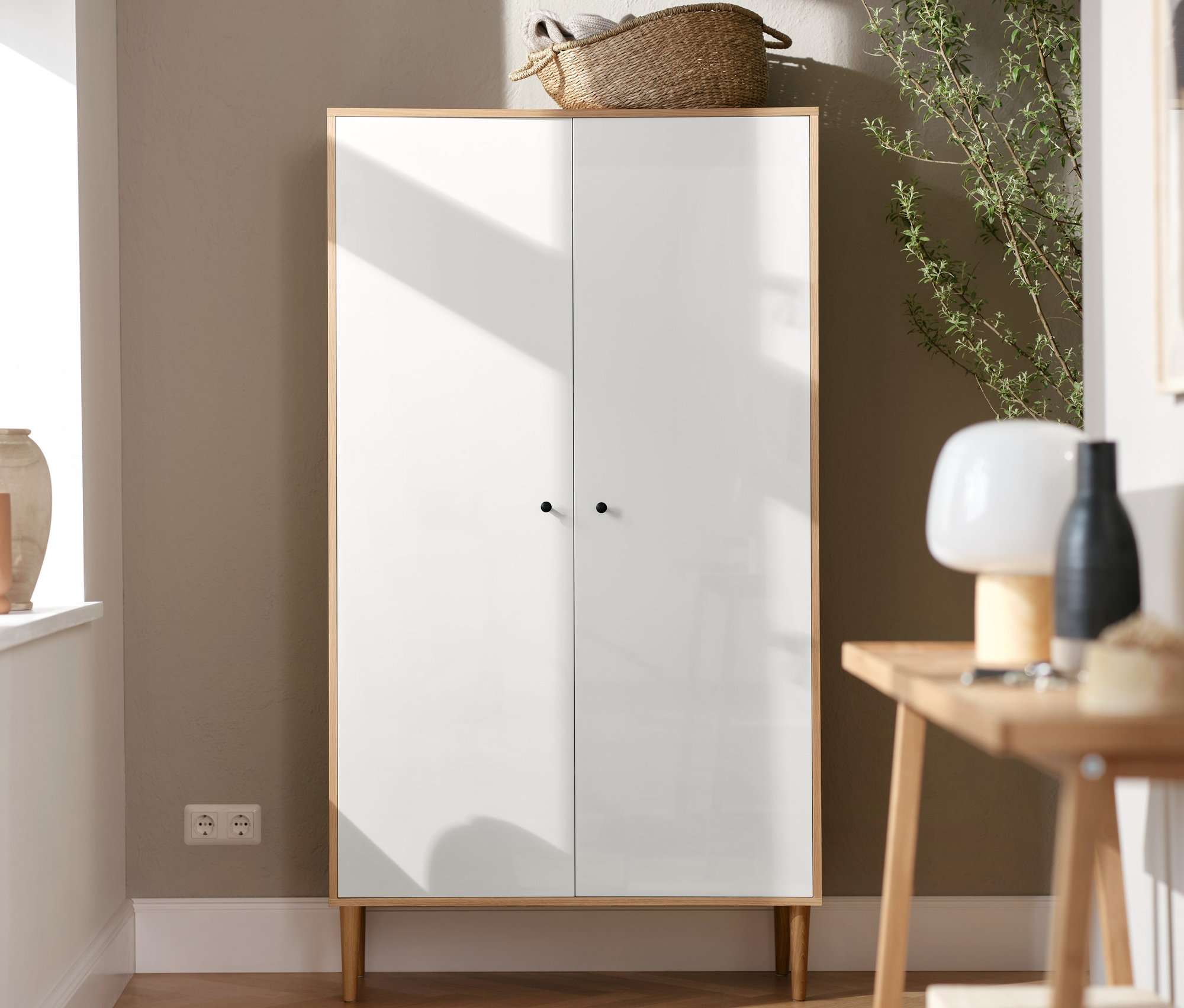 Bílá skříň stojí vedle konzolového stolku se 2 odkládacími plochami. Na stolku stojí LED akumulátorová stolní lampa.