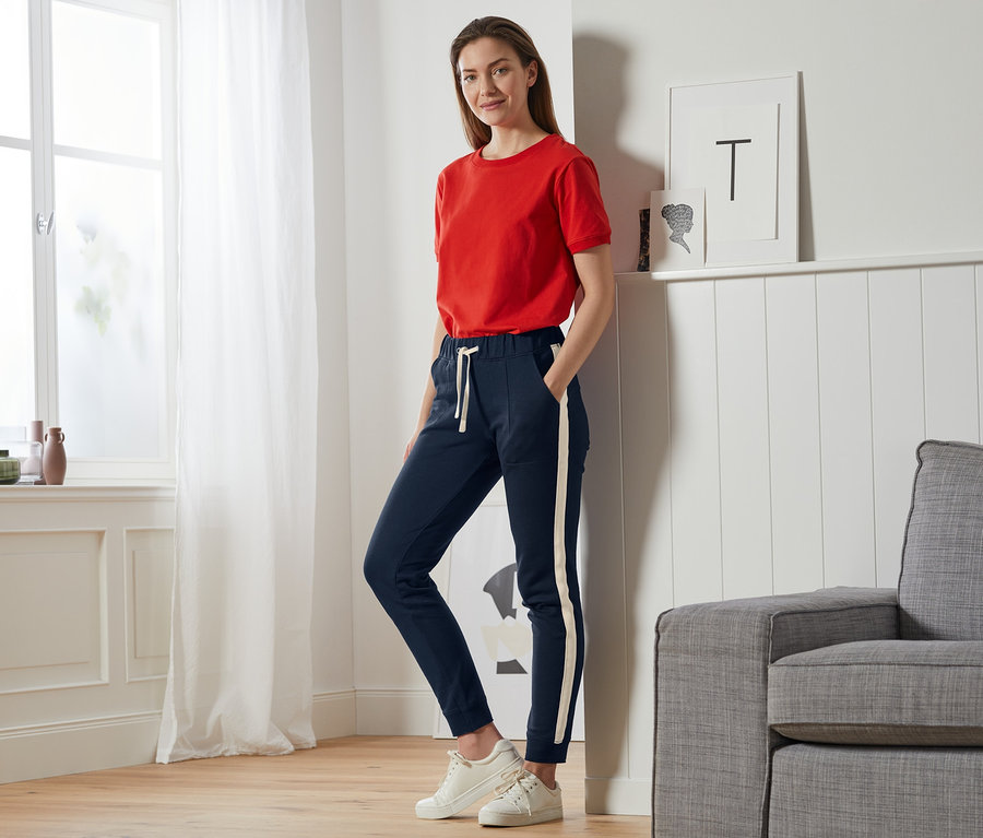 Žena stojí v obývacím pokoji a má na sobě červené tričko a tmavě modré pohodlné kalhoty.