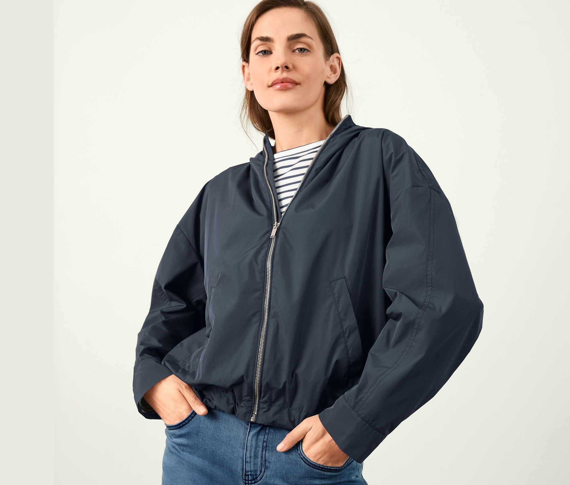 Žena pózuje v krátké parke s kapucí a v modrých džínách.