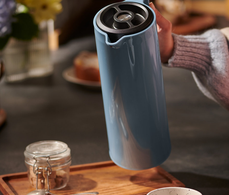 Ruka drží modrou termosku nad šálkem a dřevěným podnosem.