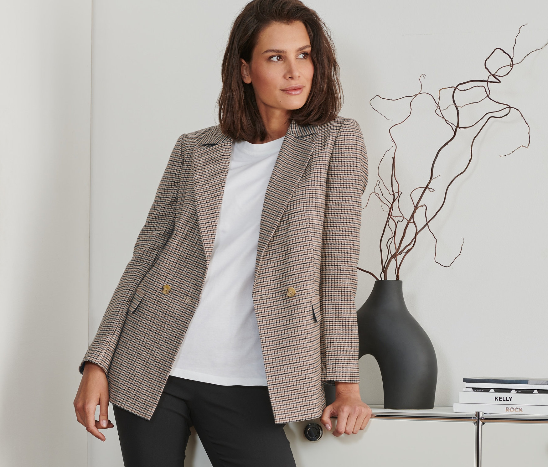 Žena opřená o bílou komodu má na sobě kostkovaný blejzr, bílé tričko a černé kalhoty.