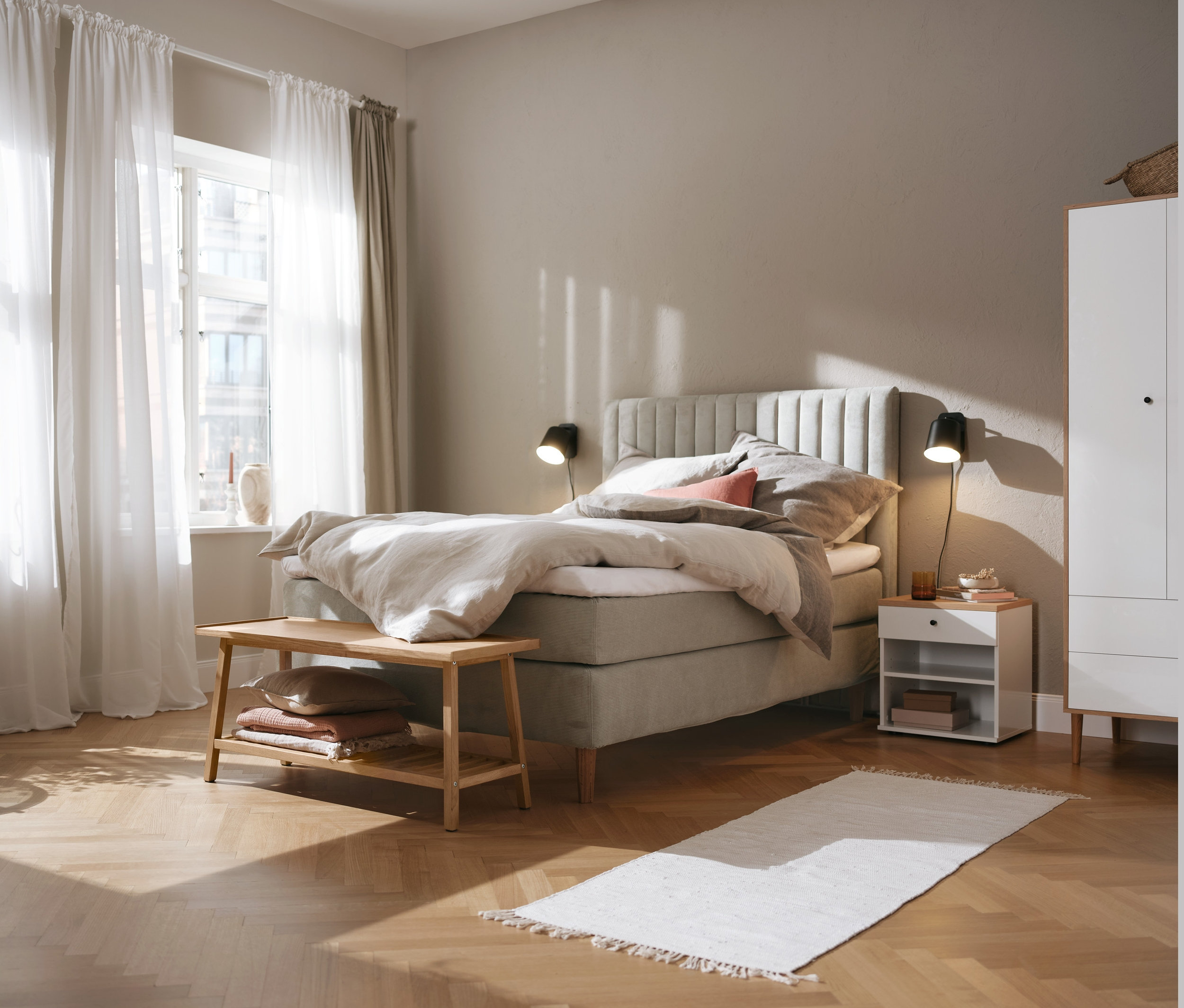 Béžová postel boxspring 180 x 200 cm, šatní skříň a noční stolek Bekeson, černé kovové nástěnné svítidlo a lavice na sezení s odkládací plochou.