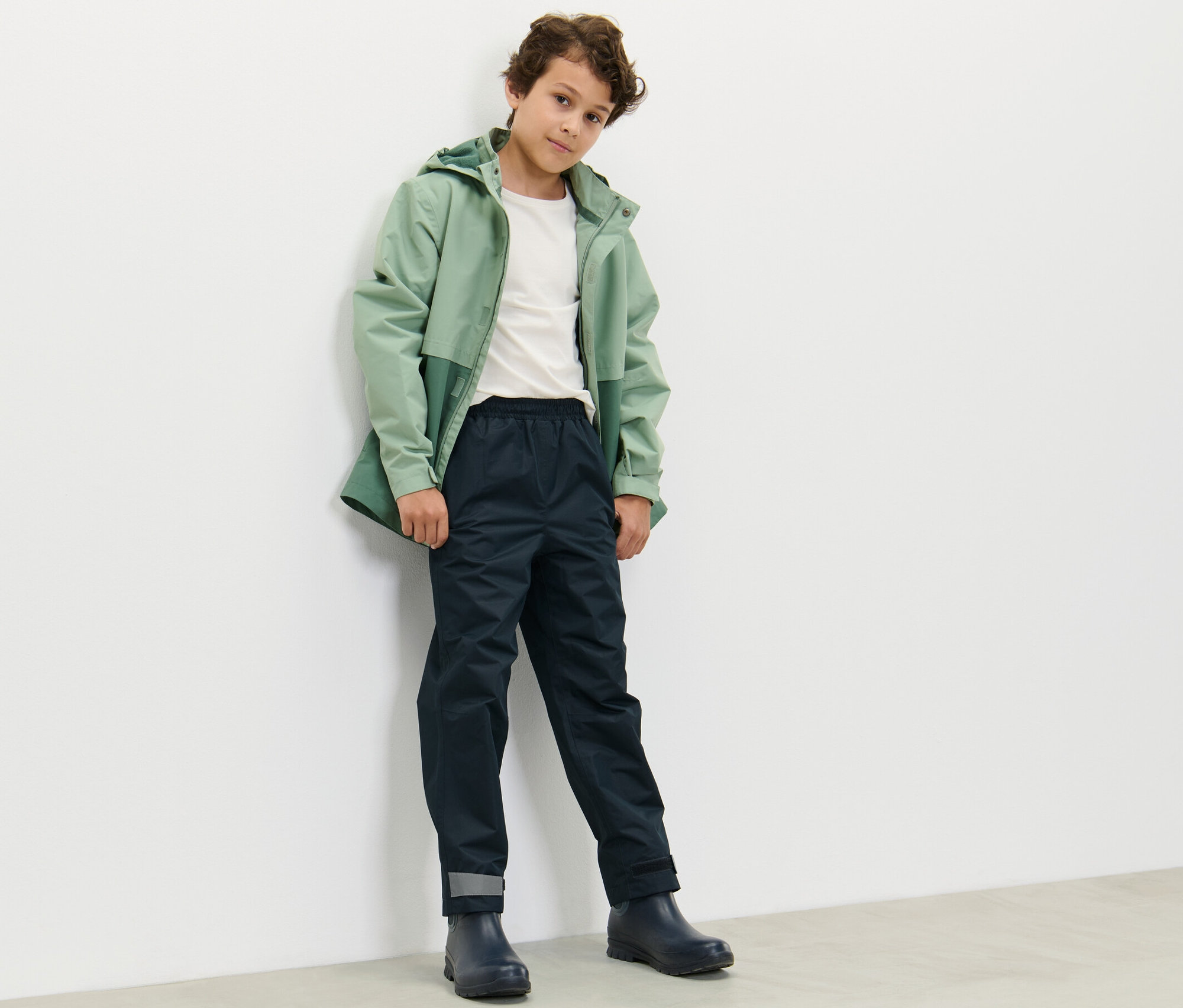Chlapec v zelené bundě a tmavě modrých dětských kalhotách do deště stojí opřený o bílou zeď.