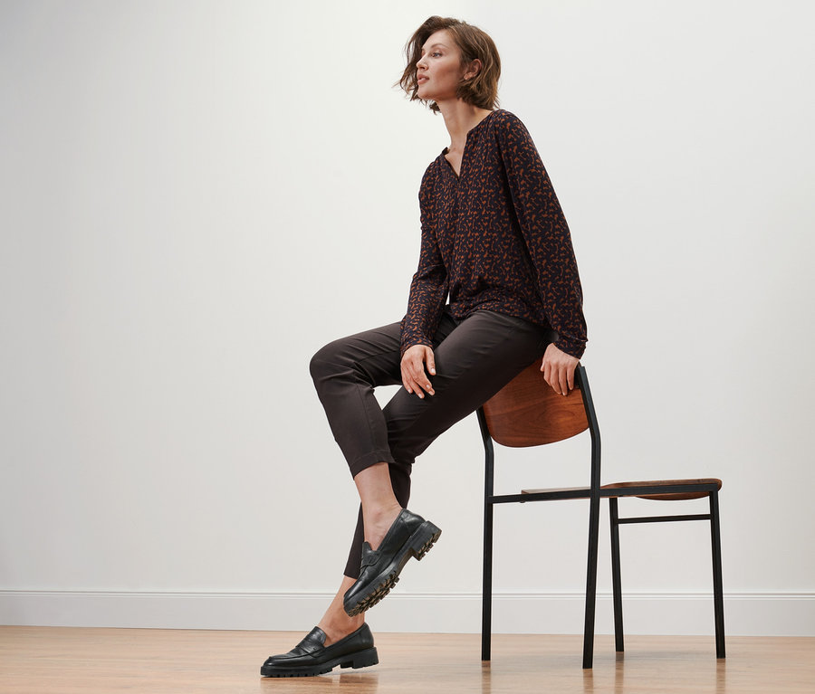 Žena sedí na židli, má na sobě kožené mokasíny, bengalinové kalhoty a halenkové triko s potiskem.