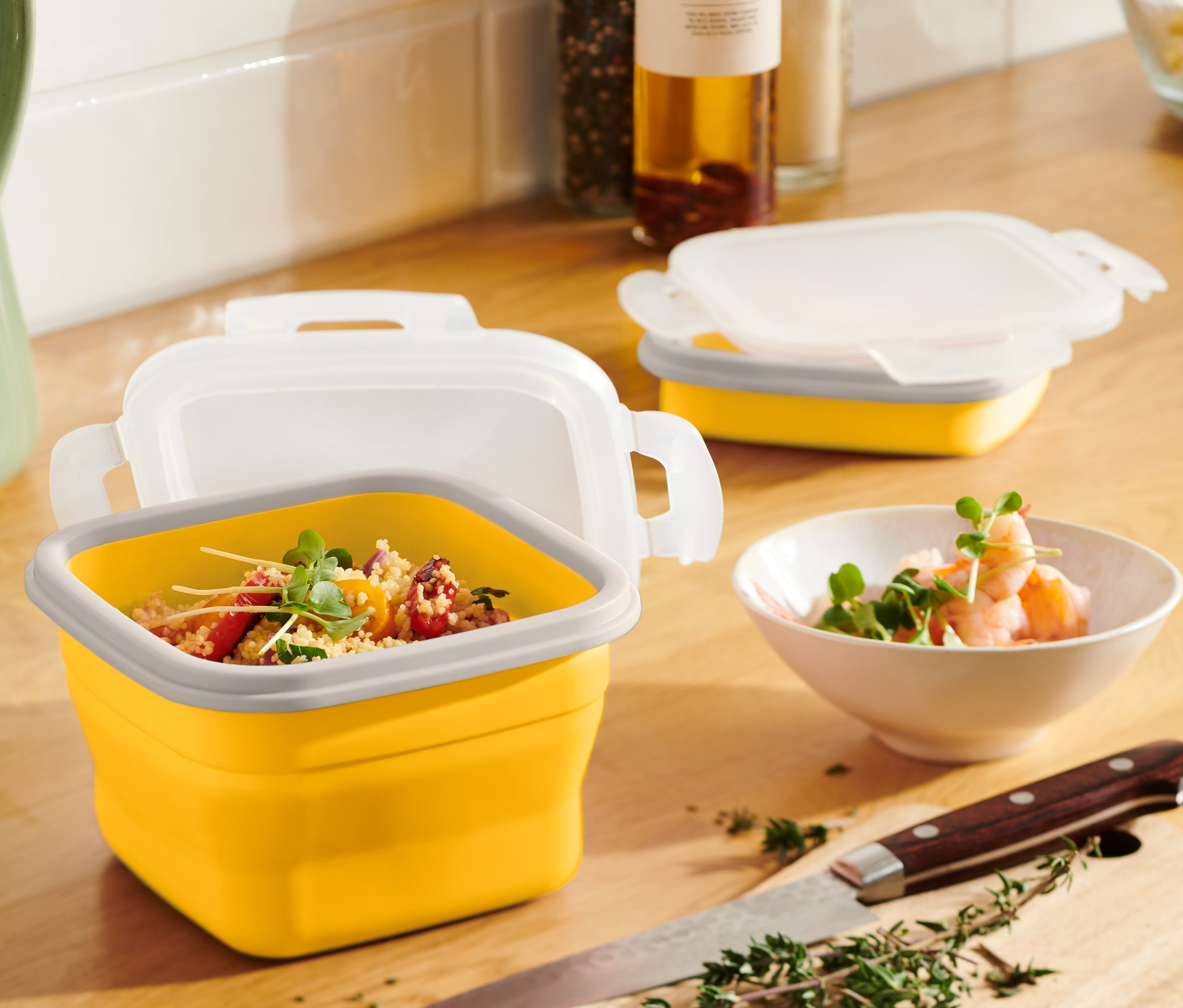 Žlutá skládací dóza na potraviny s obsahem stojí na dřevěném stole vedle zeleného džbánu a mísy s krevetami.