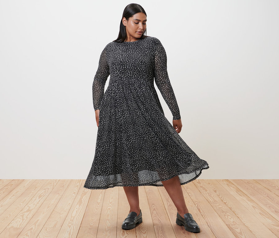 Žena v puntíkovaných síťovaných šatech a kožených mokasínech stojí na dřevěné podlaze.