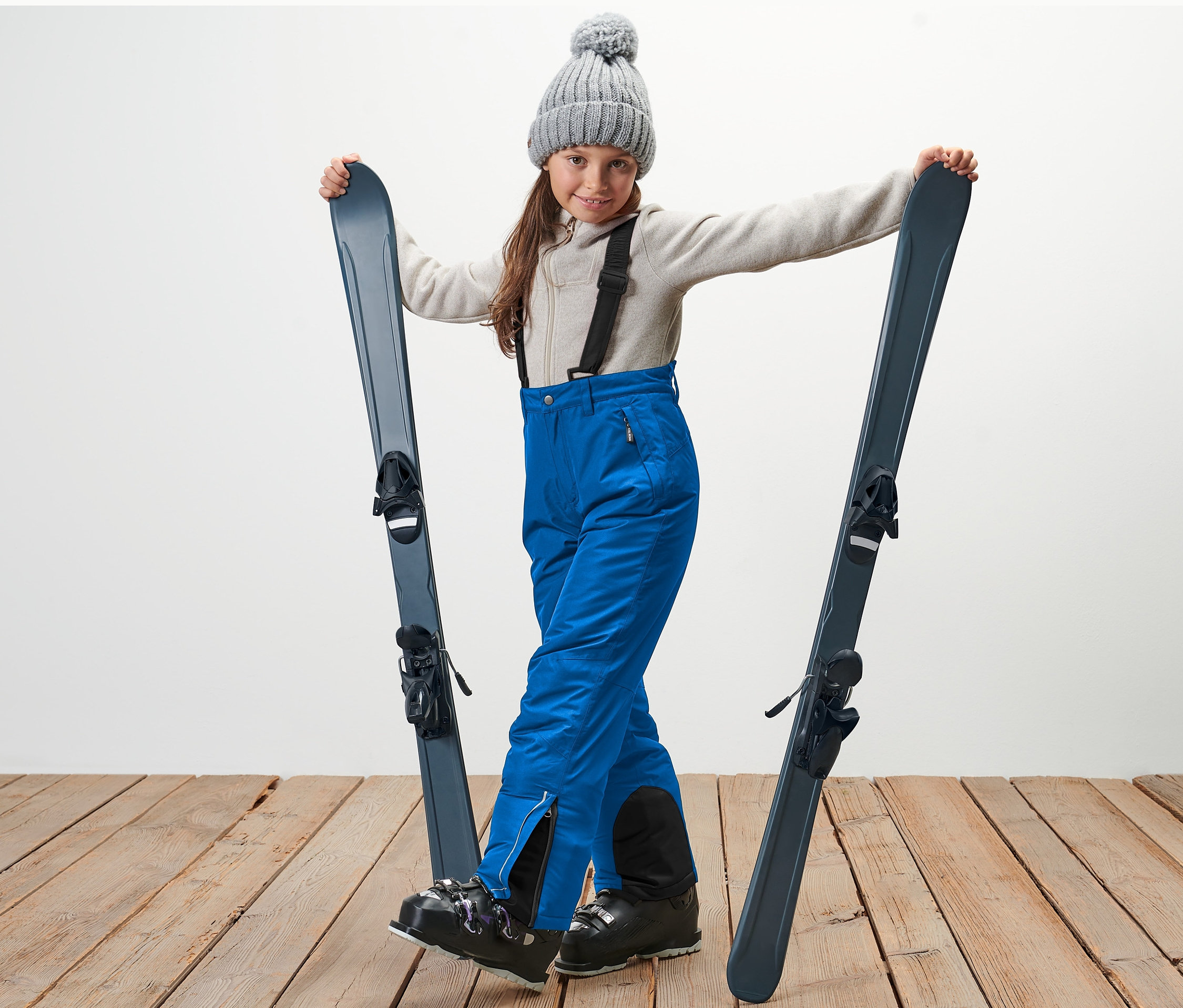 Dívka v šedé čepici drží lyže a má na sobě modré dětské lyžařské kalhoty.