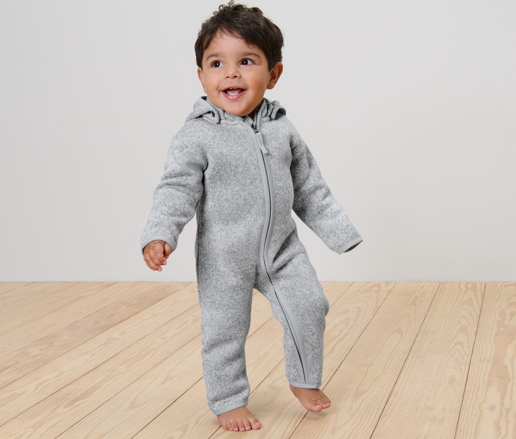 Usmívající se dítě stojí na dřevěné podlaze a má na sobě šedou dětskou kombinézu z pleteného fleecu.