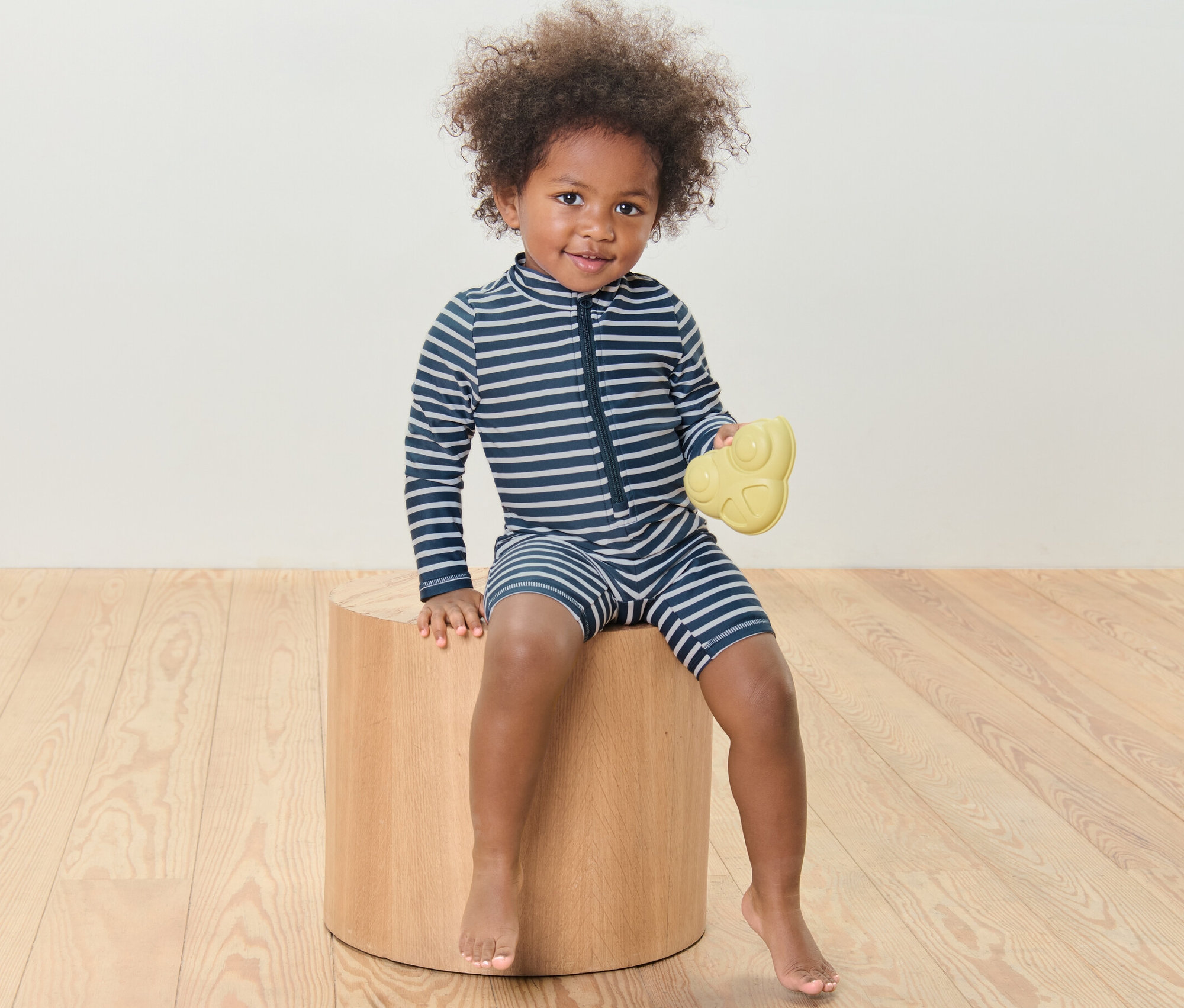 Dítě sedí na dřevěné stoličce a drží žlutou hračku, má na sobě overal s UV ochranou 80.