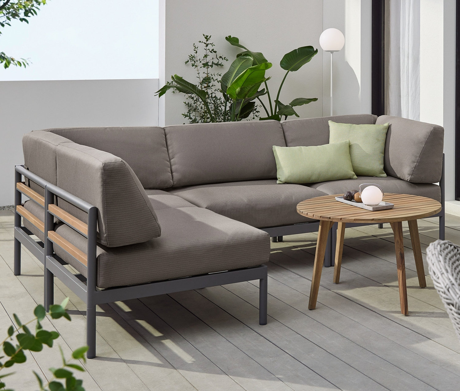 Rohová sedací souprava s šedým čalouněním Premium, kulatý konferenční stolek a odkládací stolek »Lenja« se 2 úrovněmi. Na stolech stojí LED akumulátorové lampy.